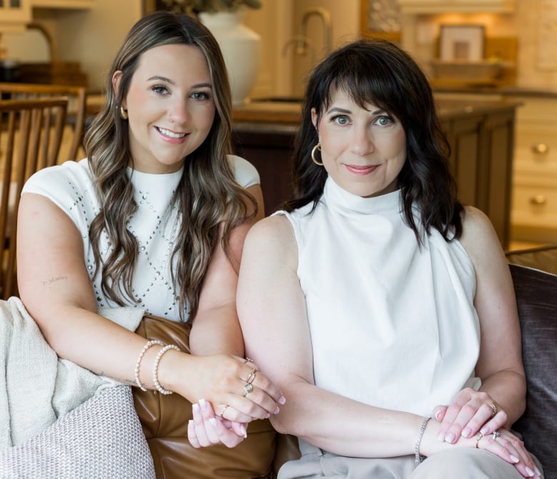 Two women smile for a photo. One has long, curling brown hair. The other has long, dark hair. They're sitting on a couch in a living room.