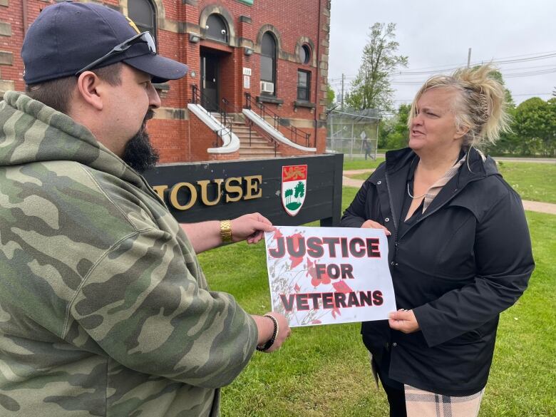 A man and woman stand in front of a brick building and hold a sign with the words, 