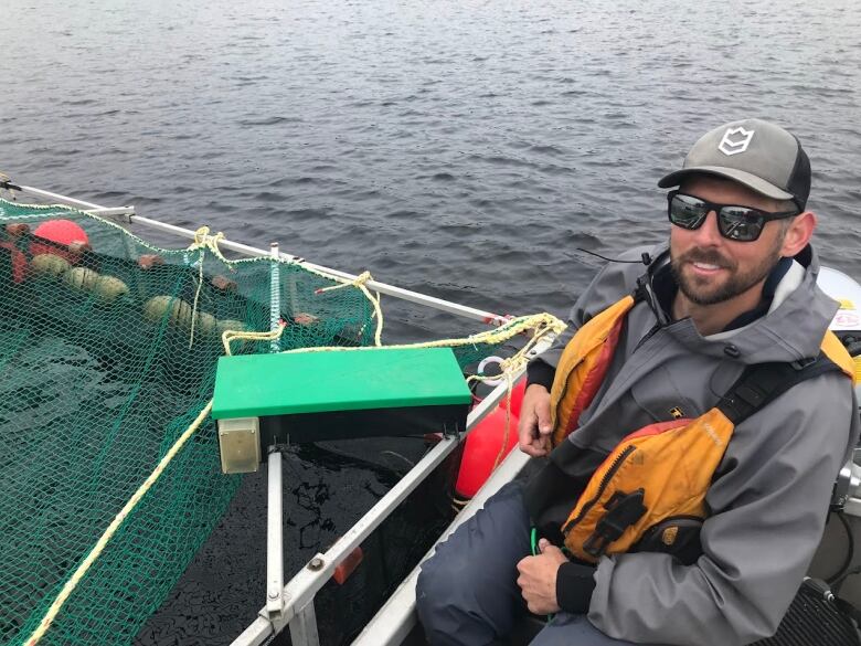 A man wearing a life jacket, a baseball cap, and a pair of sunglasses, sits on a boat. A net and rope can be seen to his left. 