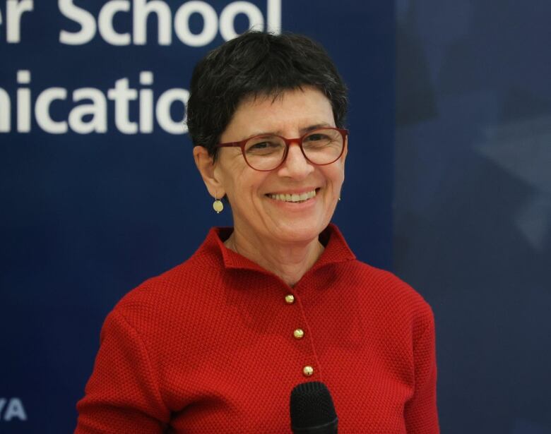 A woman with short dark hair, wearing a read sweater and red glasses, photographed in front of a blue background. 