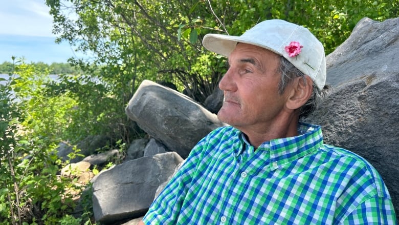 A man in a bright teal and blue plaid shirt. Looks out pensively to the river. He has a pink flower broached to his hat. 