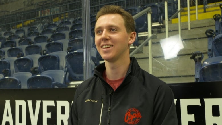 A man wearing a black jacket sitting on a hockey bench inside an arena. 