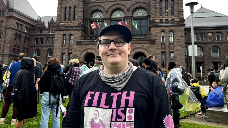 A trans person wearing a baseball cap stand in front of Queen's Park.