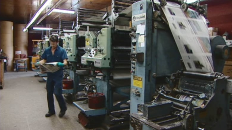 A man stands and inspects a newspaper next to a printing press