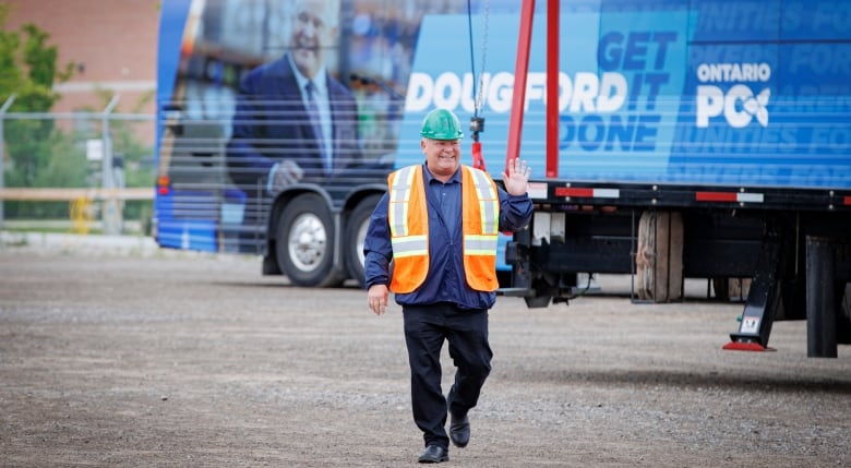 Doug Ford, wearing a safety helmet and reflective orange vest, waves as he walks in front of the blue Ontario PC Party election campaign bus with the slogan 'Get It Done.'