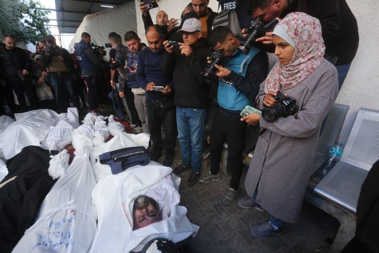 A crowd of people holding cameras look down at a the body of a man lying on the ground wrapped in a white body bag, next to other body bags. 