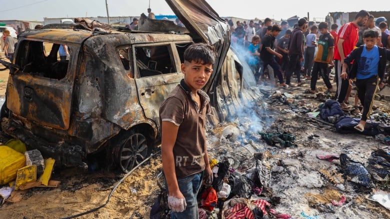 A boy stands in front of the wreckage of a burned vehicle destroyed in an airstrike, with ash and burned clothing strewn on the ground, while a crowd of people examines the scene in the background. 