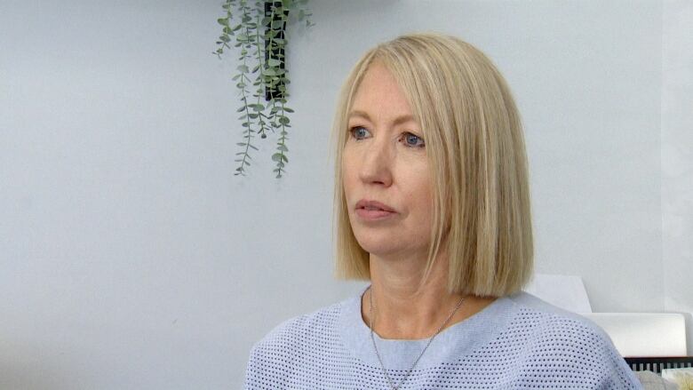 A white woman with a blonde bob and light purple sweater sits in a white office with a green plant trailing down beside her head