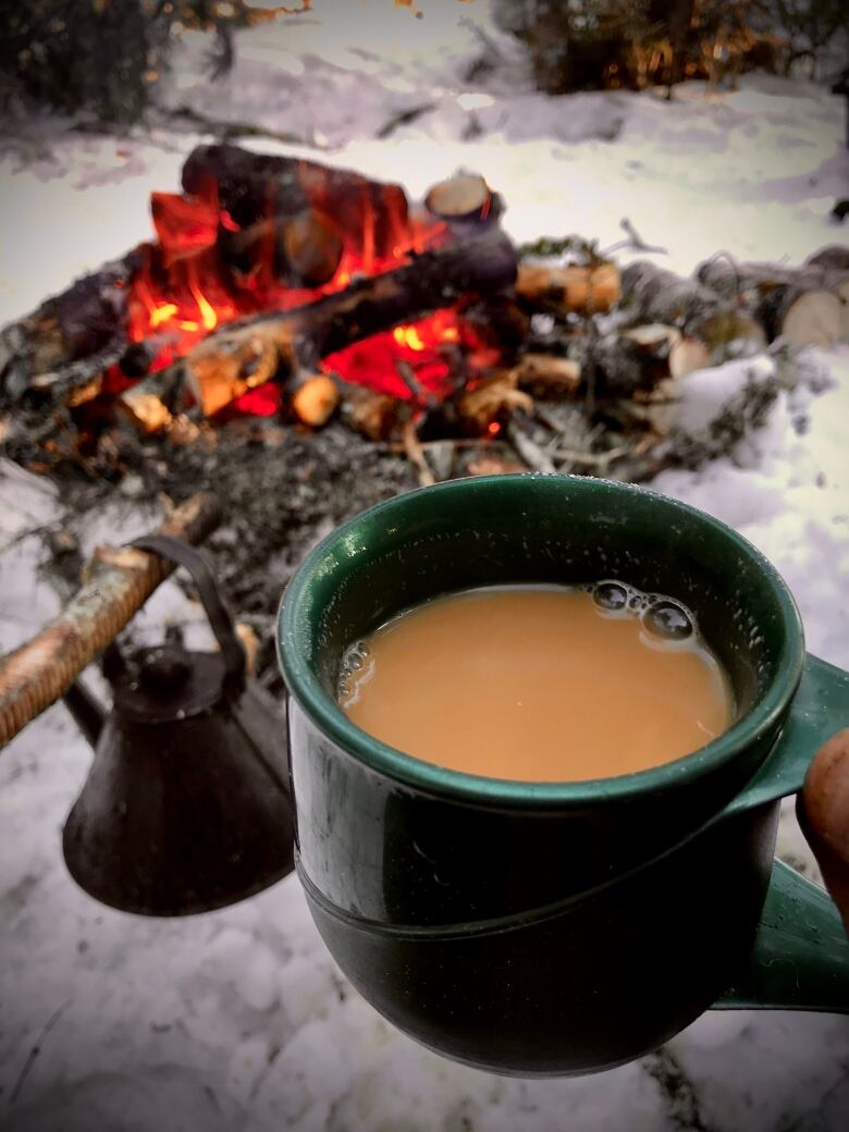 A hand holds a cup of tea with milk above a campfire in the woods.