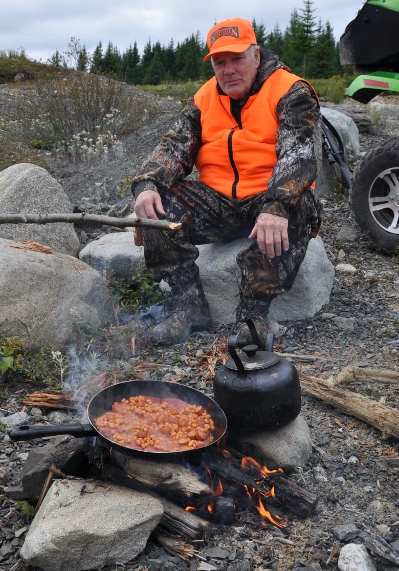 A man in camouflage outerwear and a bright orange vest sits on a large rock in front of a campfire. A frying pan on the fire is cooking food.