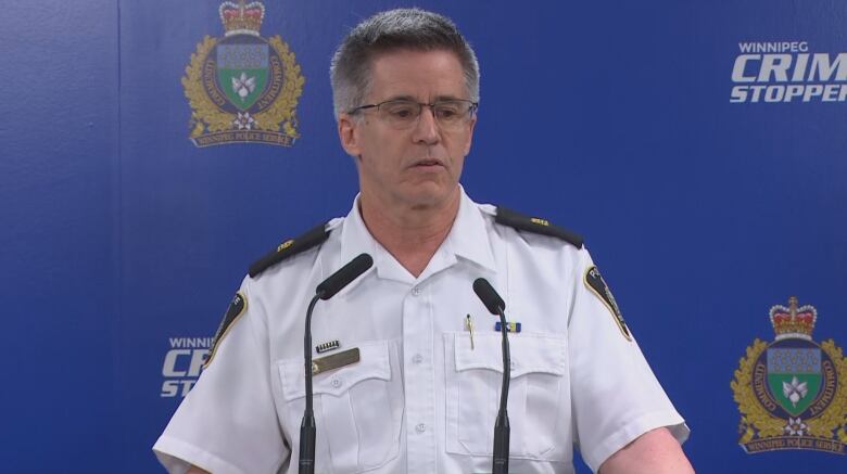 A man in a white collared shirt and short grey hair stands at a podium with microphones.