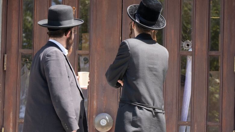 Two men have their backs to the camera. They look at a hole in a door.