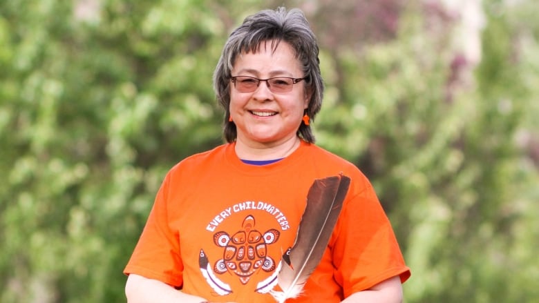 A woman wearing sunglasses smiles at the camera. She is wearing an orange shirt that says the words 'Every Child Matters.'