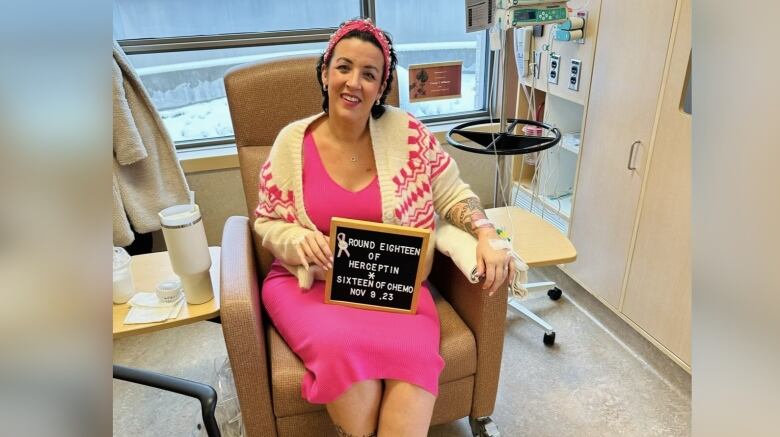 A woman wearing a hot pink dress sits on a hospital chair holding a sign that reads, 