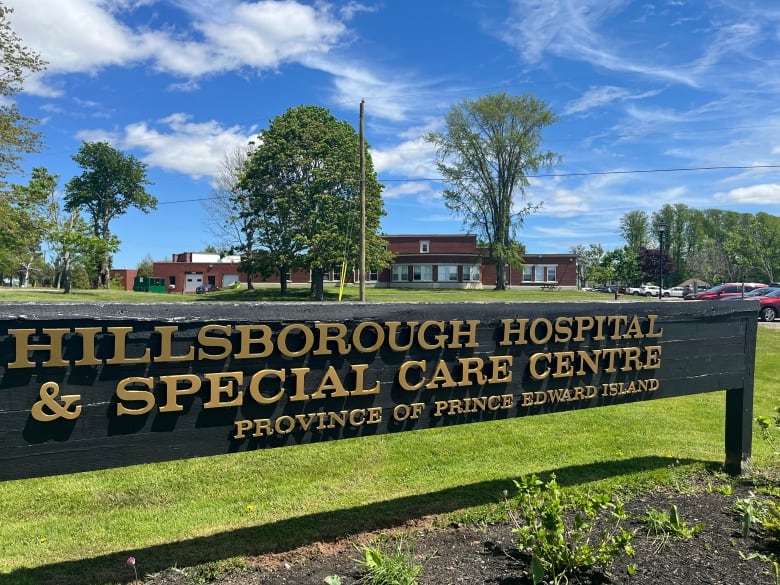 A black sign with gold letters sits in front of a brick hospital on a bright sunny, blue sky day. 