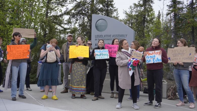 Protesters stand with signs by legislative assembly sign. 