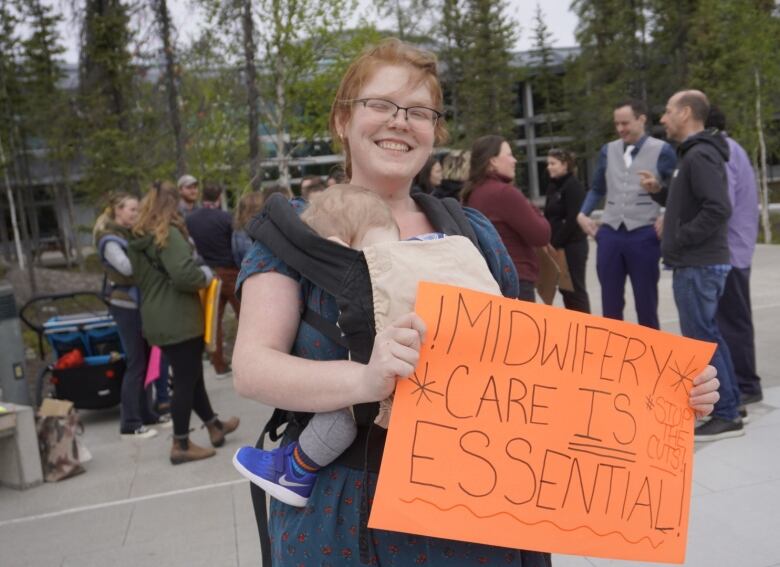 Woman with baby holds sign at protest. Sign reads, 'midwifery care is essential!'