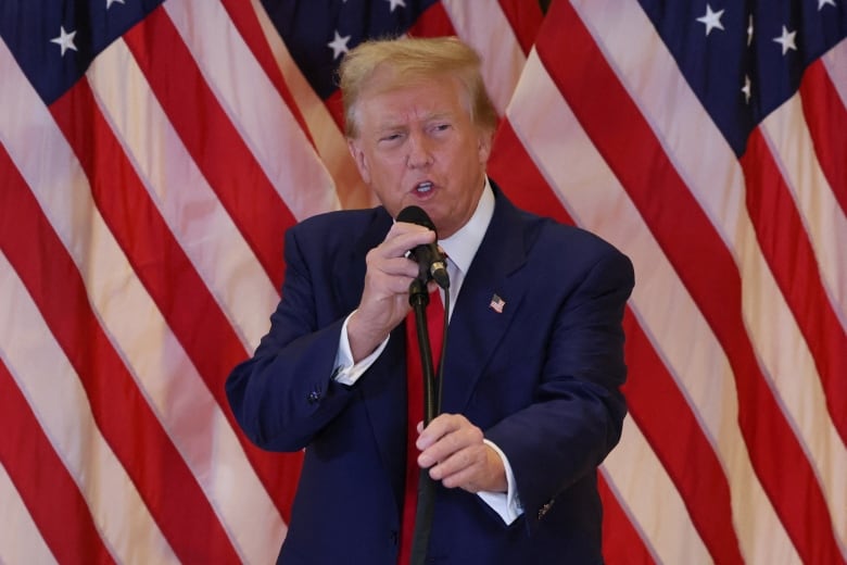 An older, clean-shaven man in a suit and tie is shown speaking before a backdrop of American flags.