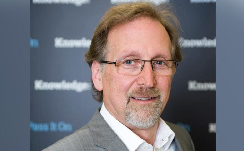 An older white man wearing glasses, a white shirt and gray blazer stands in front of a blurred out background. 