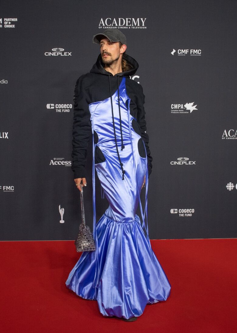 Vincent Dufault on the Canadian Screen Awards red carpet wearing a purple, floor-length dress over a black hooded sweatshirt with a black baseball cap. 