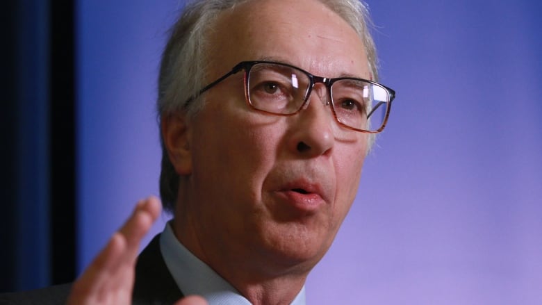 Portrait of B.C. Conservative Party Leader John Rustad speaking appears in the foreground with an out-of-focus background which looks like a purple screen 