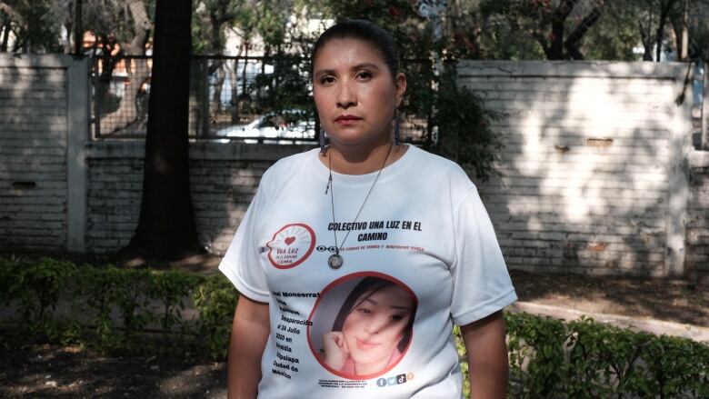 A woman with black hair, wearing a white T-shirt with slogans on it, stands in a park.