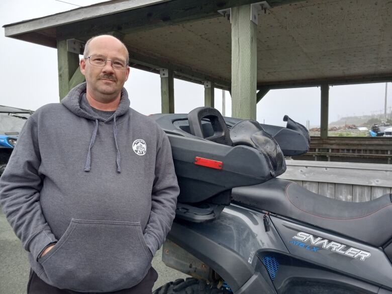 Man with mustache in a grey hoodie standing next to all terrain vehicle. A gazebo is in the background.