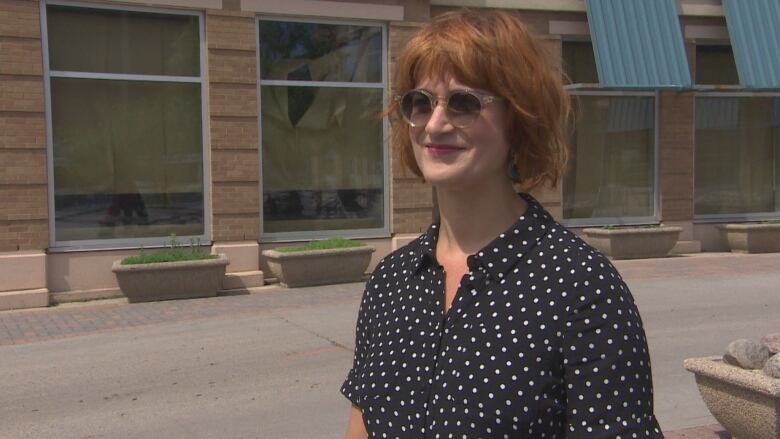 A woman in a polkadot dress, red hair and sunglasses stands outside