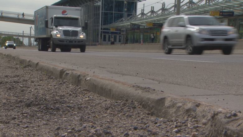 The curbs and gutters along a major roadway are worn down and crumbling. Cars are driving along the road beside it.