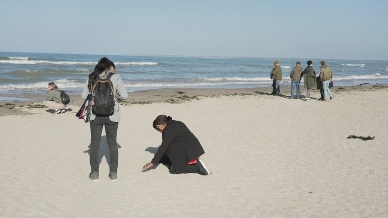 Two women crouch on a beach and touch the sand.