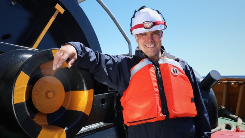 A smiling man in a hard hat and bright orange lifejacket stands on the deck of a ship on a sunny day.