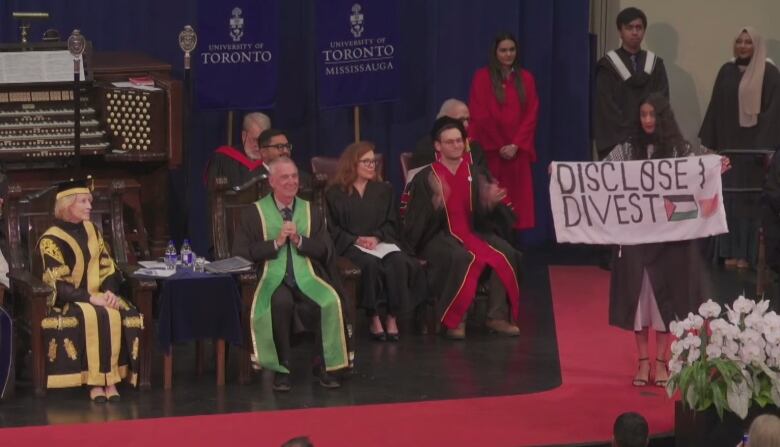 A person on stage graduating holding a banner that says 'disclose, divest'.