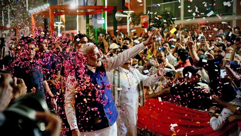 A bearded man waves outdoors to a large crowd as petals of flowers and confetti float in the air.