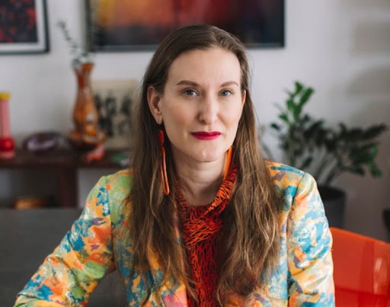 A woman with long brown hair, red lipstick and a patterned jacket and shirt sits at a counter with her arm leaning against it, posing for a photo.