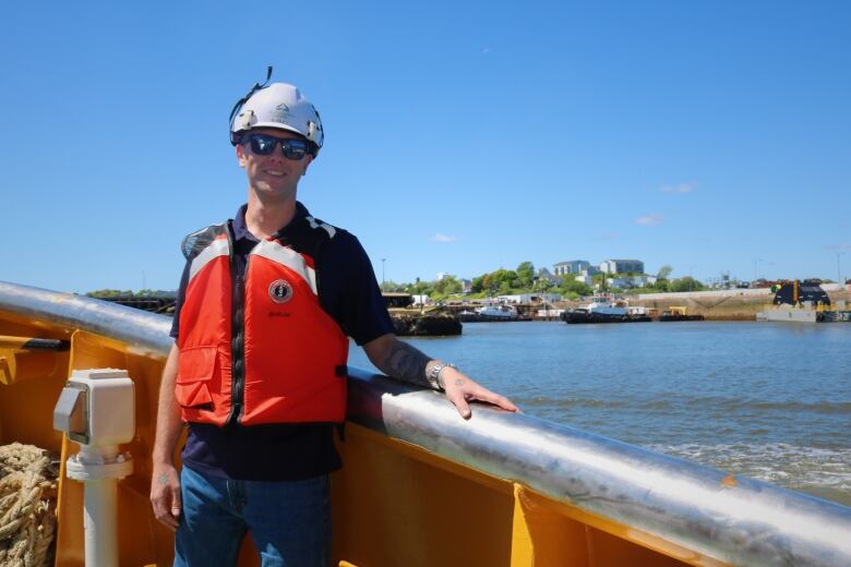 A smiling young man in an orange lifejacket stands on the deck of a tugboat overlooking the west side of Saint John. 