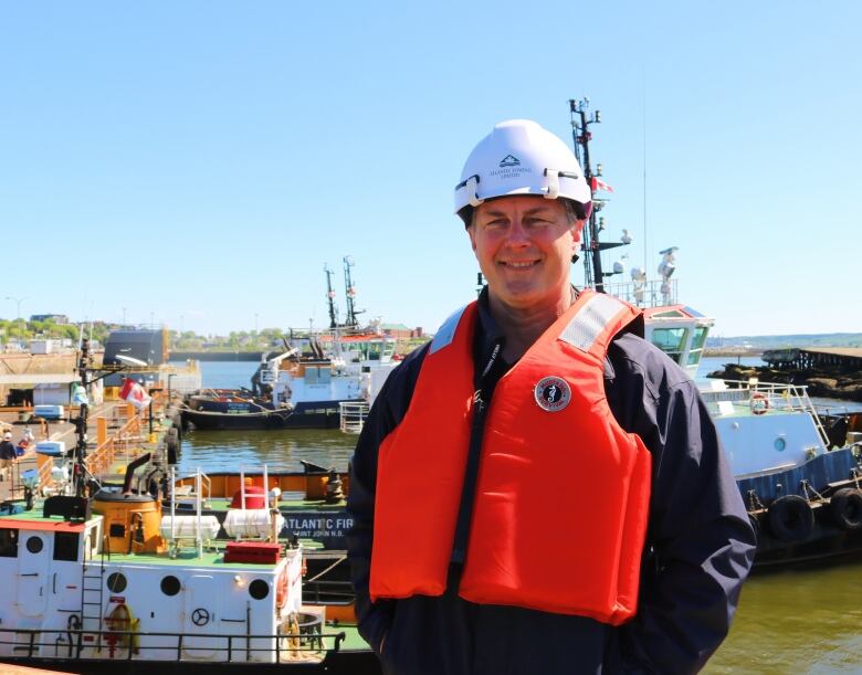 A man in a helmet and a life vest stands in front of a pier where several tugboats are moored. 