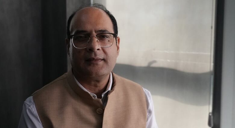 A balding man wearing glasses and a tan-coloured vest sits near a window.