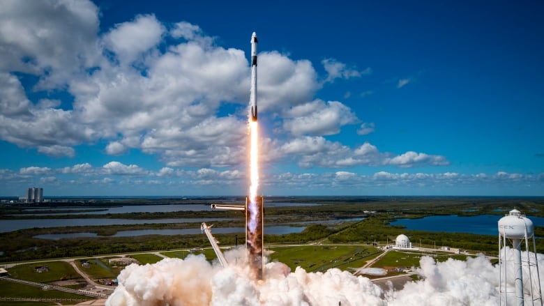 A white rocket leaves a trail of smoke on a launch pad as it soars into a blue sky dotted by white clouds.