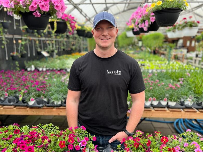 A man wearing a baseball cap and black t-shirt stands in a row of flowers at a garden centre. 