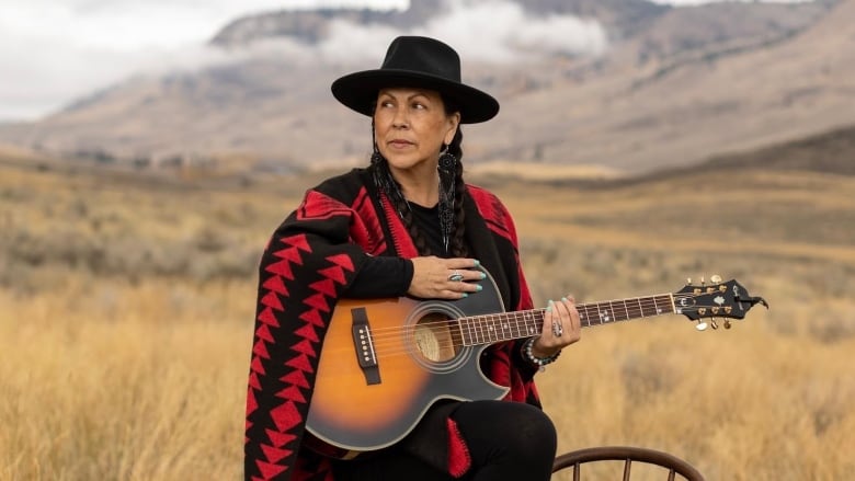 A First Nations woman wearing a cape with red designs holds a guitar, with her foot on a chair, as she gazes out over a field of dry grasses. In the background, there are mountains and clouds. 