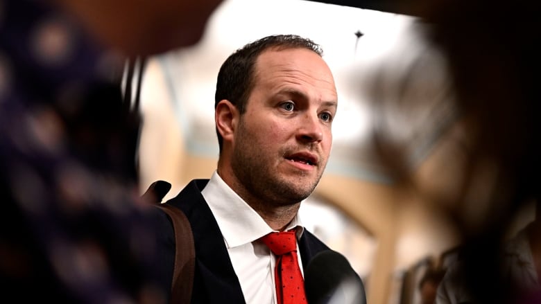 A man in a suit and red tie speaks to people not shown in the photo.