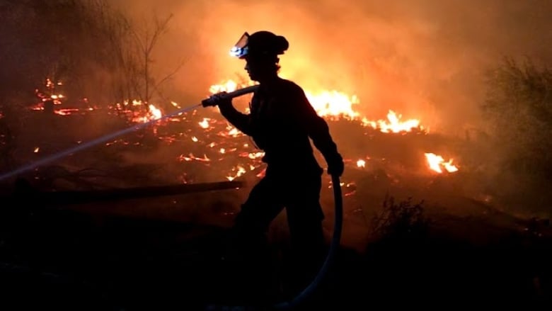 At night, a woman holds a hose over her shoulder as water streams from it. Flames are smoke are visible in the background.