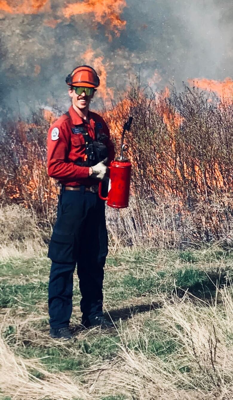 A man in a red shirt smiles as he holds a red canister with a long nozzle. The brush behind him is on fire.