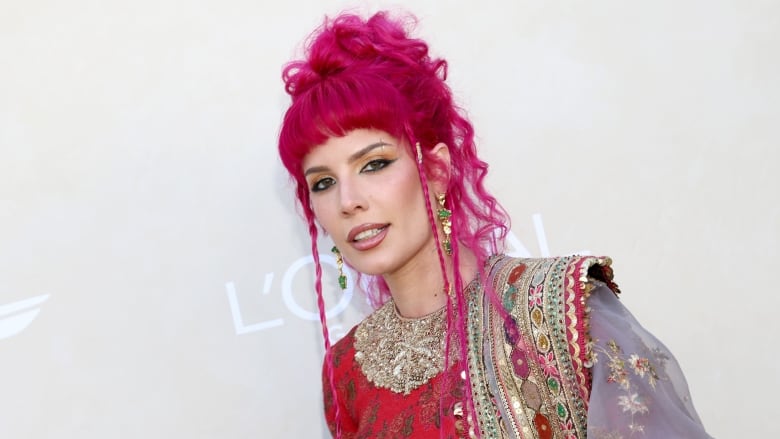 A woman with pink hair stands in front of a photo wall. 