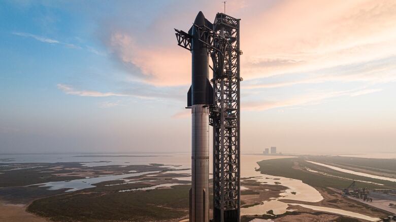 A tall rocket sits on a launch pad on a cloudy day surrounded by sand, some green grass and water.