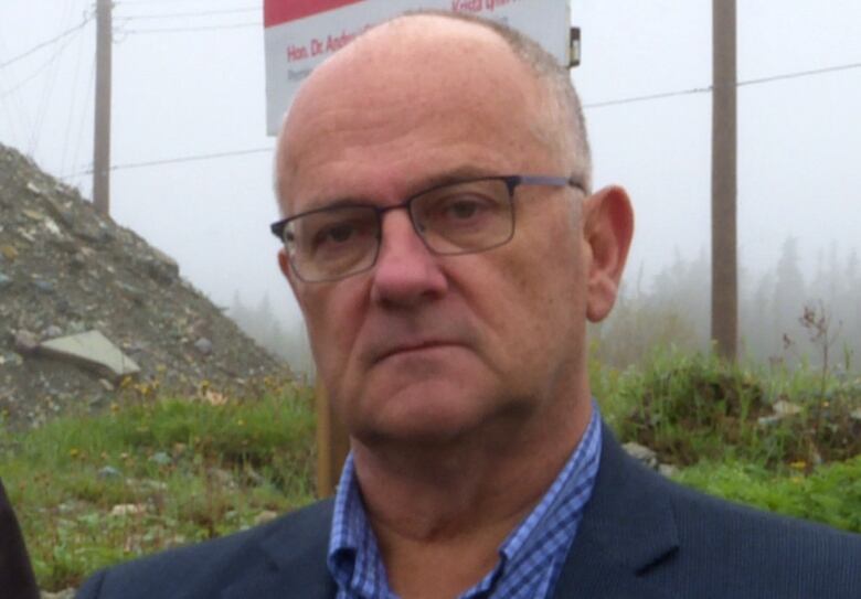 Man in suit stands in front of another man with grey hair with a pile of gravel behind them.