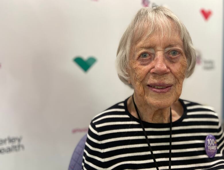 A 100-year-old woman in a black and white striped sweater poses with a purple pin on her shirt that reads 