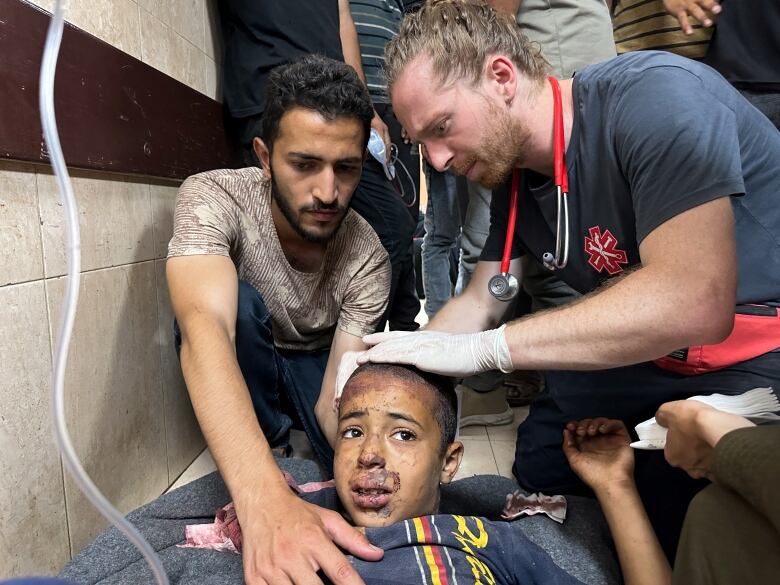 Two men, one wearing gloves tend to a small boy with a worried look and debris on his face.