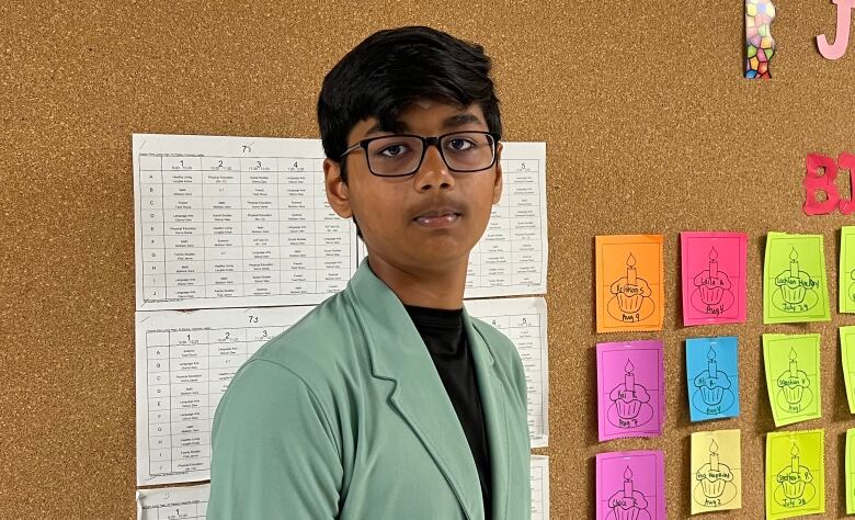 Boy in green blazer and glasses stands near cork board.