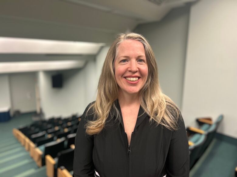 A smiling woman with long, blonde hair stands in a lecture theatre.